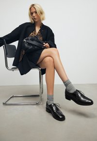 Scarpe oxford nere in pelle con dettagli brogue, indossate con calzini grigi, abbinate a un cardigan nero sopra una maglietta con stampa leopardata e una gonna nera.