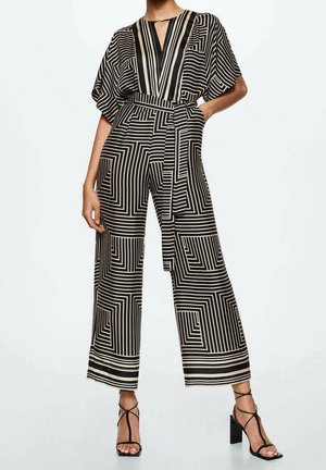 Frau in einem weit geschnittenen Jumpsuit mit schwarz-weißem geometrischem Muster, V-Ausschnitt und Gürtel, kombiniert mit schwarzen, schnüren High Heels.