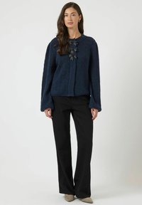 Cardigan blu navy con trama, maniche lunghe, chiusura frontale con bottoni e tre fiocchi neri sul petto. Abbinato a pantaloni neri a gamba larga.