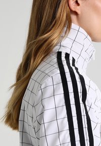 Blond kvinna som bär en vit jacka med svarta rutlinjer och tre vertikala svarta ränder på axeln, vänd åt höger.