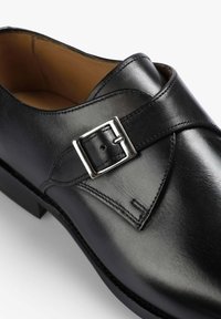 Zapato de vestir de cuero negro con una correa de hebilla cuadrada plateada y interior marrón claro, mostrado en primer plano sobre un fondo blanco.
