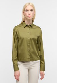 Camicia button-up verde oliva a maniche lunghe realizzata in un tessuto liscio e lucido. Presenta un colletto, bottoni e polsini; lunghezza crop.