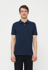 Uomo in piedi davanti a uno sfondo uniforme, indossa una polo blu navy a maniche corte e pantaloni neri, guardando avanti con postura rilassata.
