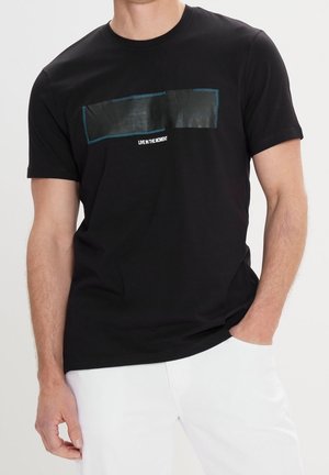 Schwarzes Baumwoll-T-Shirt mit einem rechteckigen schwarzen Stoffpatch, das mit blauen Nähten und weißem Text "LIVE IN THE MOMENT" versehen ist. Verstellbare Passform.