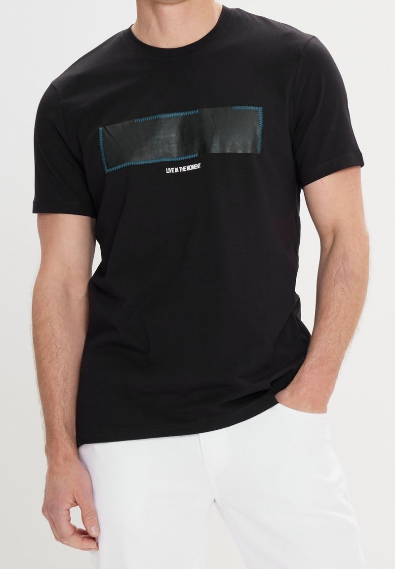 Camiseta de algodón negra con un parche rectangular de tela negra con costuras azules y texto blanco que dice "VIVE EN EL MOMENTO." Ajuste regulable.