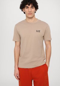 Beige bavlněné tričko s krátkým rukávem. Obsahuje malé černé logo EA7 na hrudi. Hladká textura, volný střih.