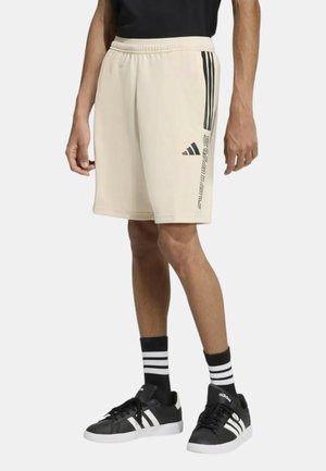Uomo che indossa pantaloncini beige Adidas con strisce nere e logo, calzini a righe bianche e nere e scarpe da ginnastica Adidas nere.