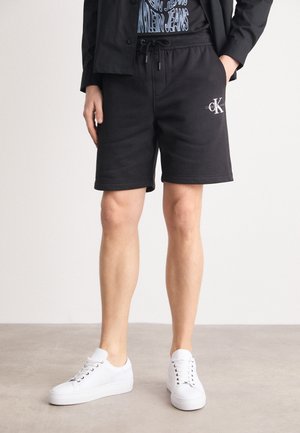 Modèle masculin portant un short noir Calvin Klein, une veste noire et des baskets blanches, debout sur un sol gris devant un fond blanc.
