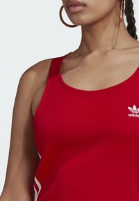 Červený bavlněný top bez rukávů se širokými ramínky a kulatým výstřihem. Na levé části hrudi je malá bílá značka adidas. Hladká textura, bez vzorů.