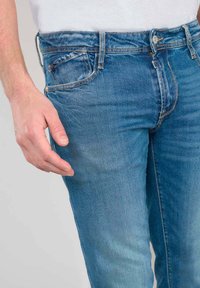 Jean en denim bleu slim fit au look délavé, avec cinq poches, des passants de ceinture et une fermeture à un bouton. Le tissu a une texture lisse.
