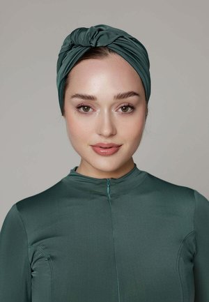 Jonge vrouw met lichte make-up die een donkergroene tulband hoofdband en bijpassende top met lange mouwen draagt, kijkt recht vooruit tegen een effen achtergrond.