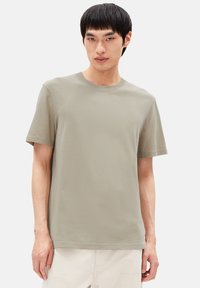 Junger Mann in einem schlichten hellolivgrünen T-Shirt mit kurzen Ärmeln und cremefarbenen Hosen, der vor einem weißen Hintergrund steht.