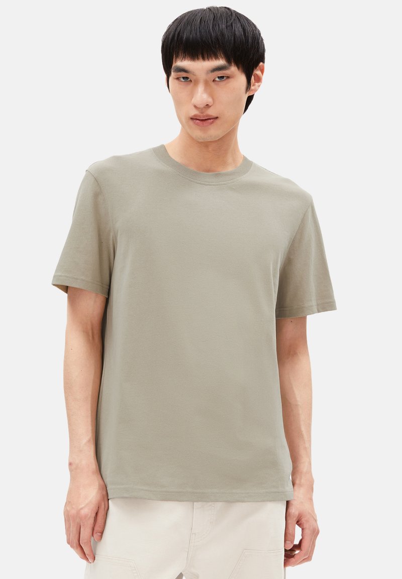 Junger Mann in einem schlichten hellolivgrünen T-Shirt mit kurzen Ärmeln und cremefarbenen Hosen, der vor einem weißen Hintergrund steht.
