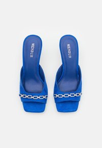 Mules de ante azul con punta abierta y una amplia correa que presenta un detalle de cadena plateada, suela plana y plantilla acolchada para mayor comodidad.