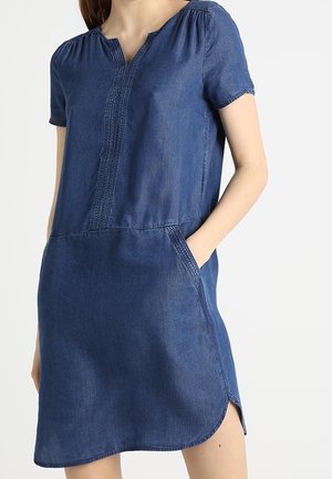 Mujer con vestido vaquero azul de manga corta, con bolsillos laterales y una pequeña abertura en el dobladillo inferior.