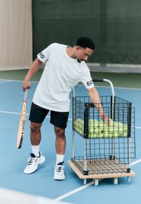 Tenniskølle, hvit kortermet skjorte, svarte shorts. Metallvogn med grønne tennisballer. Blå tennisbane, hvite sokker og sportssko.