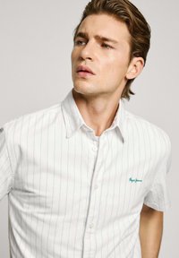Camisa de botones de manga corta blanca con rayas verticales azul claro, con un cuello casual y un logo bordado verde en el pecho.