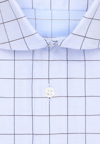Camicetta a quadri azzurri chiari con colletto aperto, caratterizzata da un piccolo bottone bianco al collo e da motivi a griglia scura su un tessuto dalla texture particolare.
