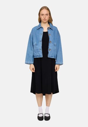 Light blue denim jacket with button closure, worn over a black dress. Black shoes and white socks complete the outfit. Front view.
