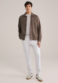 Camicia marrone con bottoni e taschino sul petto, indossata sopra una maglia grigia chiara, abbinata a pantaloni bianchi e sneaker grigie con dettagli marroni.