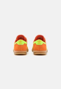 Idrottsskor med en ljus orange mockaövre, neongröna detaljer och en genomskinlig gummisula. Har logotyp vid hälen och mönstrad mesh.