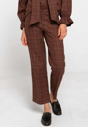 Piernas vistiendo pantalones de cuadros marrones de pierna ancha combinados con mocasines de cuero negros, manos relajadas a los lados, de pie contra un fondo liso.