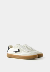 White low-top sneakers with gum soles, black chevron detail on sides, white laces, and padded collars, shown from a side angle.