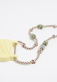 Funda de teléfono de silicona amarilla unida a un cordón texturizado trenzado de colores púrpura y crema, con nudos verdes y un broche plateado.