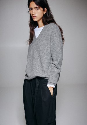 Un mannequin pose devant un fond de studio gris, vêtu d’un pull gris ample sur une chemise blanche et d’un pantalon noir.
