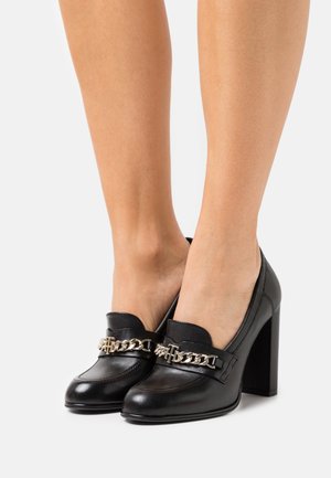 Loafers à talons en cuir noir avec un détail en chaîne sur le devant, dotés d'un bout arrondi et d'une surface texturée, avec un talon carré rehaussé.