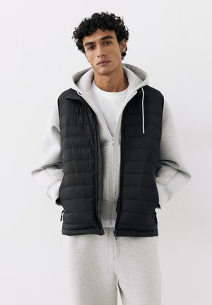 Jeune homme aux cheveux bouclés portant un sweat à capuche gris et un pantalon de survêtement, superposé d'un gilet matelassé noir, debout devant un fond blanc.