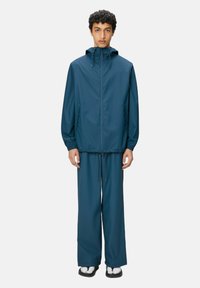 Giacca a vento con cappuccio e pantaloni abbinati in verde scuro. Presenta polsini elasticizzati, tasche laterali e cintura regolabile con cordino.