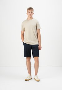 Beige pamut póló zsebbel, sötétkék rövidnadrággal párosítva. A modell krémszínű sportcipőt visel, fehér háttér előtt áll.