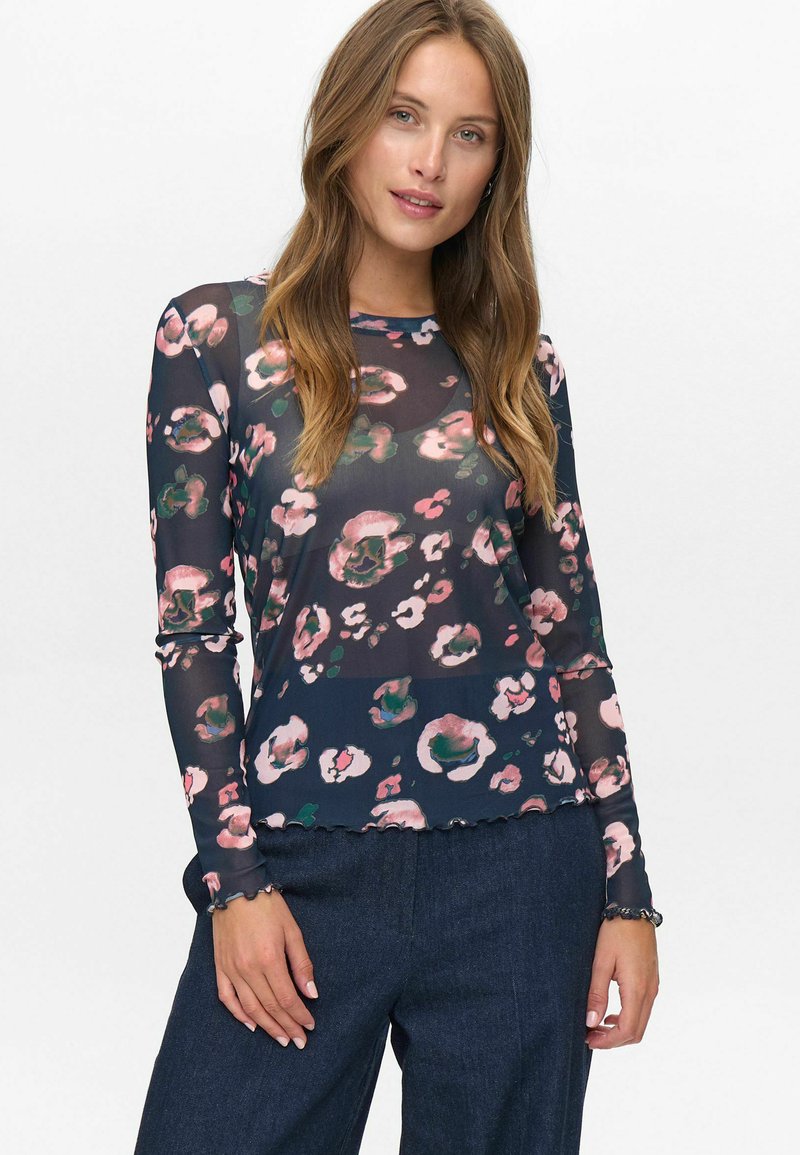 Donna che indossa una blusa lunga a maniche trasparenti di colore blu navy con stampa floreale rosa e verde, abbinata a pantaloni larghi blu scuro, in piedi su uno sfondo bianco.