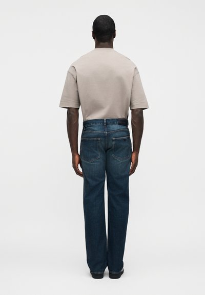 Χαλαρή γκρι βαμβακερή μπλούζα, σε συνδυασμό με σκούρα μπλε τζιν παντελόνια με ίσια γραμμή, που διαθέτουν κλασικές πίσω τσέπες και εμφανείς ραφές.