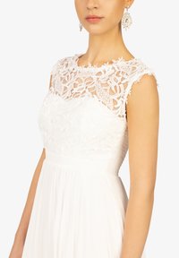 Robe blanche avec un corsage en dentelle à motifs floraux, une taille cintrée et une jupe fluide. Complétée par des boucles d'oreilles pendantes.