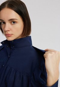 Mornaroplasty dugmadna košulja s visokim ovratnikom, nabrana sprijeda i s kopčama na dugme. Tkanina izgleda glatko s lagano strukturiranim dizajnom.