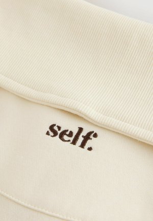 Cream-colored fabric with a ribbed collar, featuring brown embroidered text reading "self." on the front. Smooth texture and minimal design.