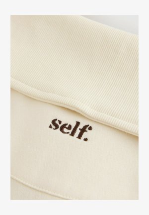 Cream-colored fabric with a ribbed collar, featuring brown embroidered text reading "self." on the front. Smooth texture and minimal design.