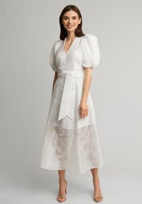 Biała sukienka z bufiastymi rękawami, głębokim dekoltem w kształcie litery V, zdobiona teksturowanym korpusem i haftem kwiatowym. Posiada pasek w talii i przezroczystą, warstwową spódnicę.