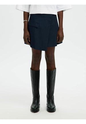 Personne portant une jupe asymétrique marine avec une poche à rabat et des bottes en cuir noires montantes, debout devant un fond uni.