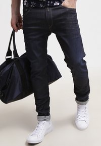 Mörka denimjeans med slim fit, uppvikta fållar och kontrasterande sömmar. Matchas med vita sneakers och en svart duffelväska.