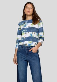 Frau, die ein blau-weiß gestreiftes Oberteil mit Sternmustern und eine blaue weit geschnittene Jeans trägt, steht mit einer Hand in der Tasche.