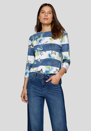 Frau, die ein blau-weiß gestreiftes Oberteil mit Sternmustern und eine blaue weit geschnittene Jeans trägt, steht mit einer Hand in der Tasche.