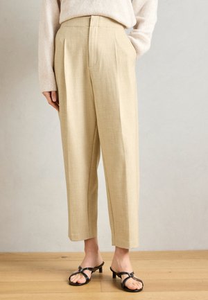 Bézs len széles szárú nadrág, elülső redőkkel, oldalzsebekkel és tapered alsó résszel, fekete magassarkú szandállal párosítva.