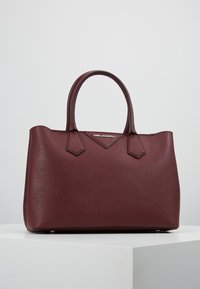 Håndtaske i burgunderfarvet syntetisk læder med to rullede håndtag, trekantede sideaccenter, glat tekstur og en sølvlogo på forsiden.