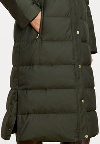 Zelený kabát s podšívkou, ktorý má prešitú textúru, zlaté kovové doplnky, bočné vrecká a mierne rozšírený spodný lem.