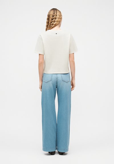 Γυναίκα με κυματιστά ξανθά μαλλιά στέκεται γυρισμένη πλάτη, φορώντας ένα απλό λευκό κοντομάνικο τοπ και ψηλόμεσα ανοιχτόχρωμα μπλε τζιν με φαρδιά μπατζάκια.