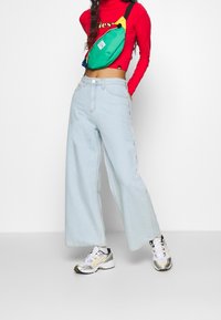 Helle blaue, weit geschnittene Jeans mit hoher Taille, kombiniert mit einem roten Langarmoberteil und einer mehrfarbigen Gürteltasche. Auffällige weiße Sneakers.