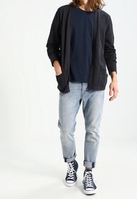 Ciemnoszary kardigan z długim rękawem, z otwartym przodem i kieszeniami, zestawiony z granatową koszulą i jasnoniebieskimi podwiniętymi jeansami, dopełniony granatowymi trampkami.